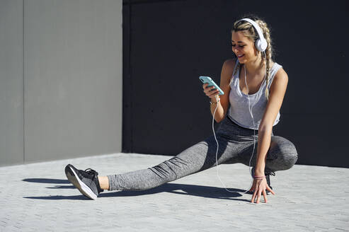 Lächelnde Sportlerin mit Kopfhörern, die ein Mobiltelefon benutzt, während sie auf dem Gehweg hockt - PGF00310