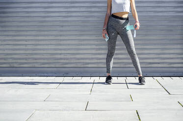 Sportswoman holding water bottle and mobile phone while standing against shutter - PGF00311