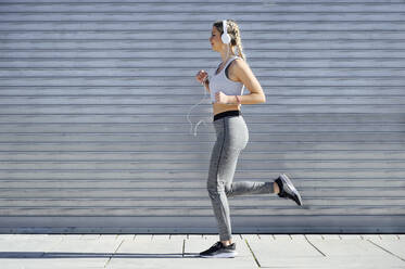 Female athlete wearing headphones jogging against shutter - PGF00312