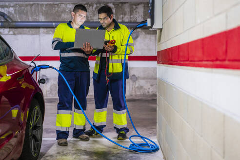 Male technicians using laptop while charging electric vehicle in auto repair shop - DLTSF01424