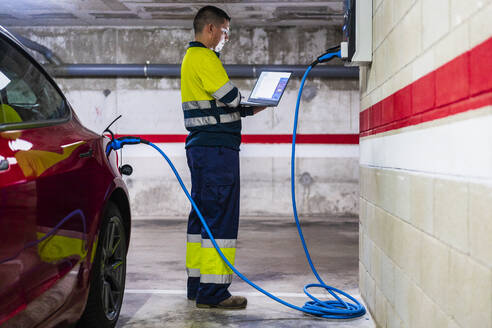 Professioneller männlicher Techniker programmiert am Laptop, während er ein Elektroauto auflädt - DLTSF01431