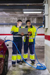 Male programmer discussing with colleague over laptop while charging electric car - DLTSF01433