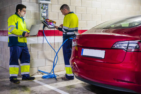 Male technicians working by meter while charging electric car in auto repair shop - DLTSF01449