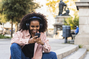 Glückliche junge Frau mit Afro, die ein Mobiltelefon benutzt, während sie auf einer Treppe in der Stadt Musik hört - PNAF00328