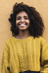 Glückliche junge Frau im Pullover an einer gelben Wand stehend - PNAF00338