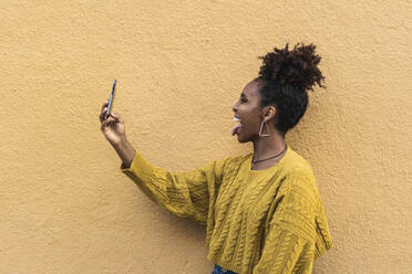 Glückliche Afro-Frau, die ihre Zunge herausstreckt, während sie ein Selfie von einer gelben Wand macht - PNAF00339