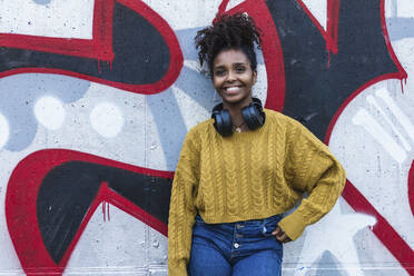 Fashionable young woman with headphones smiling while standing against painted wall - PNAF00341