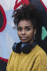 Selbstbewusste junge Frau mit lockigem Haar und Kopfhörern vor einer Graffiti-Wand - PNAF00343