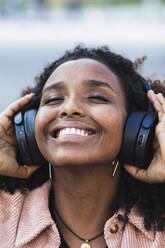Glückliche Afro-Frau mit geschlossenen Augen hört Musik über Kopfhörer in der Stadt - PNAF00351