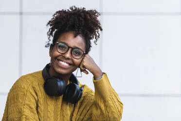 Fröhliche afrofarbene junge Frau im Pullover mit Kopfhörern vor weißer Wand - PNAF00355