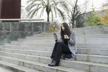 Female entrepreneur with reusable coffee cup using mobile phone on staircase - MTBF00808