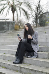 Fashionable businesswoman using mobile phone while having coffee on staircase - MTBF00809