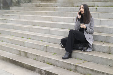 Unternehmerin mit wiederverwendbarem Kaffeebecher beim Telefonieren auf der Treppe sitzend - MTBF00810