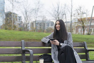 Lächelnde weibliche Fachkraft mit Kaffeetasse, die wegschaut, während sie auf einer Parkbank ihr Handy hält - MTBF00811