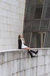 Fashionable businesswoman looking up while sitting on retaining wall of office building - MTBF00814