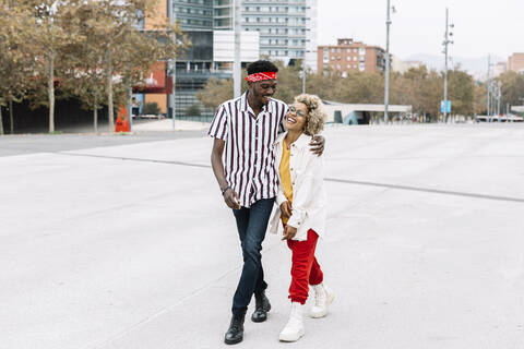 Smiling man walking with arm around female friend on footpath in city stock photo