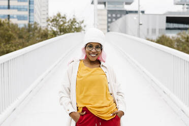 Lächelnde Frau mit rosa Perücke und Mütze steht mit den Händen in den Taschen auf einer Brücke - XLGF00930