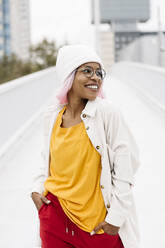 Frau mit Mütze und Perücke schaut weg, während sie mit den Händen in den Taschen auf einer Brücke steht - XLGF00931