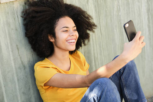Lächelnder Student, der ein Selfie mit seinem Handy macht, während er an der Wand sitzt - AODF00016