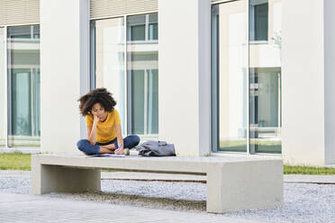 Studentin schreibt in Buch, während sie auf einer Bank auf dem Universitätsgelände sitzt - AODF00022