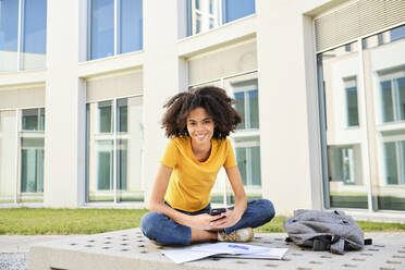 Lächelnde Studentin mit Büchern, die ein Mobiltelefon benutzt, während sie auf dem Universitätsgelände sitzt - AODF00024
