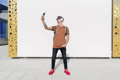 Smiling man wearing headphones taking selfie through mobile phone while standing with hands in pockets against wall stock photo