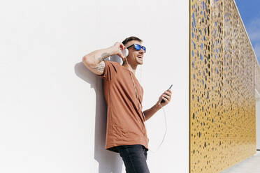 Glücklicher Mann mit Mobiltelefon, der über Kopfhörer Musik hört, während er sich an die Wand lehnt - MARF00050