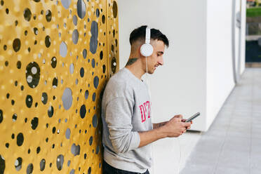 Junger Mann mit Kopfhörern, der ein Mobiltelefon benutzt und sich an eine Wand lehnt - MARF00056