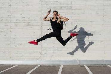 Carefree young man with mobile phone and headphones carrying backpack while jumping against wall - MARF00060