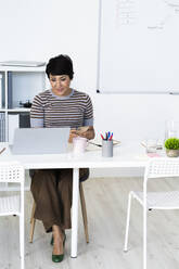 Portrait of businesswoman sitting at office table with smart phone in hands - GIOF10105