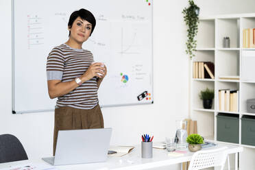 Portrait of businesswoman posing in office with mug in hands - GIOF10112