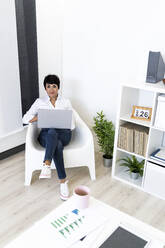 Portrait of businesswoman smiling at camera while sitting with laptop in office armchair - GIOF10168