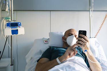 Patient mit Infusionstropf, der ein Mobiltelefon benutzt, während er im Krankenhaus im Bett liegt - JCMF01679