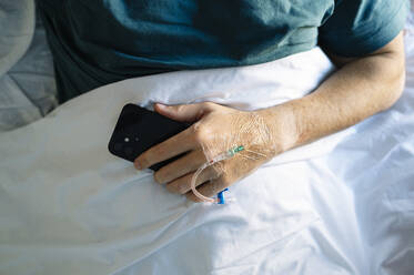 Patient hält Mobiltelefon mit IV-Tropf Hand auf dem Bett im Krankenhaus liegend - JCMF01683