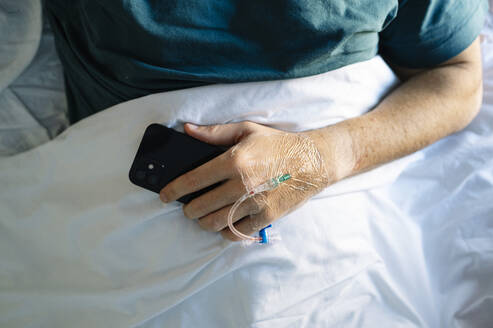 Patient holding mobile phone with IV drip hand lying on bed in hospital - JCMF01683