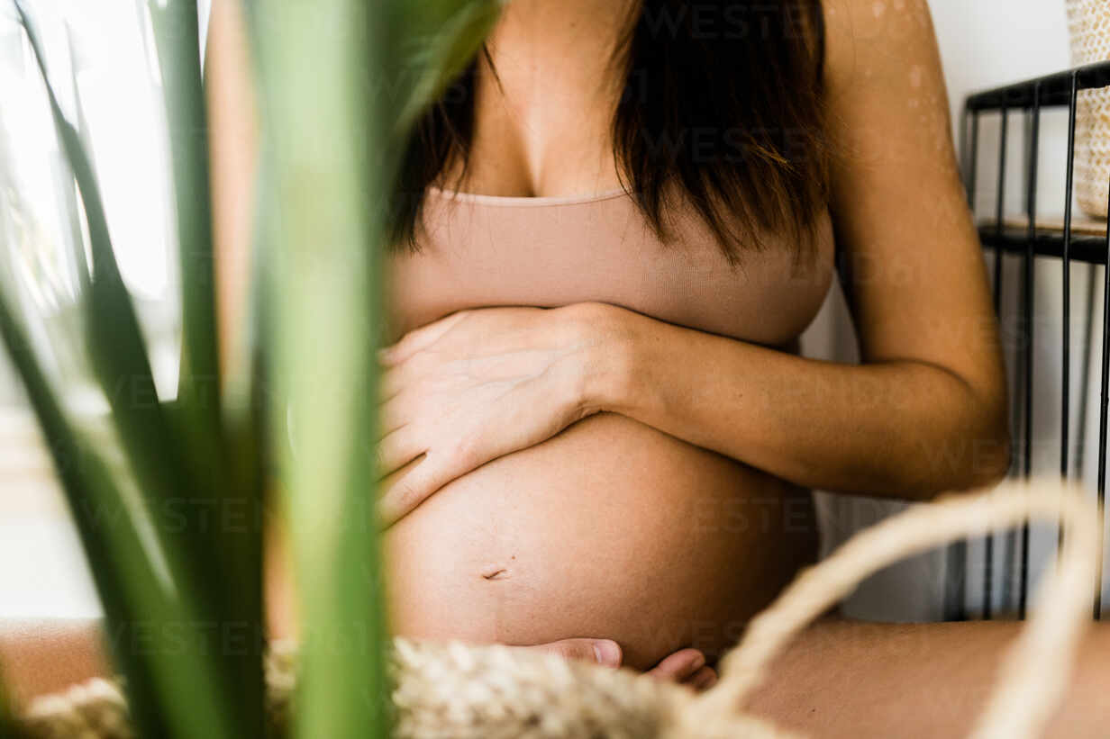 Side view of cropped serene unrecognizable pregnant female in underwear  sitting on stool in room with green houseplants and tenderly touching tummy  stock photo