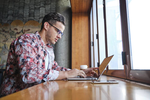 Junger fokussierter männlicher Freiberufler mit Brille und Einwegglas mit Heißgetränk auf der Fensterbank sitzend mit Netbook - ADSF19243