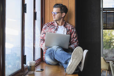 Junge trendige Fernarbeiterin mit Brille und Netbook sitzt mit gekreuzten Beinen auf der Fensterbank in der Nähe eines Einwegglases mit Kaffee und schaut aus dem Fenster - ADSF19244