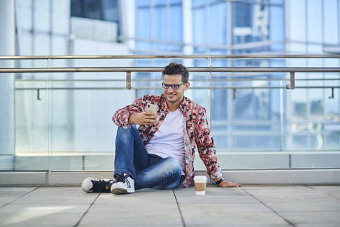 Junger lächelnder Mann in modischer Kleidung und mit Brille, der mit gekreuzten Beinen auf dem Boden in der Nähe einer Glaswand sitzt und Kaffee zum Mitnehmen trinkt - ADSF19245