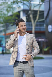Junger, fröhlicher Mann in modischer Kleidung, der in der Stadt mit dem Handy telefoniert und dabei wegschaut - ADSF19246