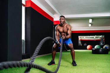 Back view of ethnic sportsman with strong torso exercising with battle ropes during functional training in gym - ADSF19259