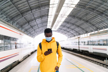 Ethnischer männlicher Reisender mit Rucksack und Maske, der auf dem Bahnsteig mit dem Zug steht und ein Mobiltelefon benutzt, während er auf die Abfahrt während der COVID 19-Epidemie wartet - ADSF19261