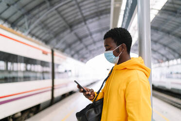 Ethnischer männlicher Reisender mit Rucksack und Maske, der auf dem Bahnsteig mit dem Zug steht und ein Mobiltelefon benutzt, während er auf die Abfahrt während der COVID 19-Epidemie wartet - ADSF19262