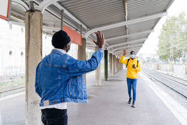 Black male friends in medical masks waving hands and greeting each other on train platform during coronavirus epidemic - ADSF19264