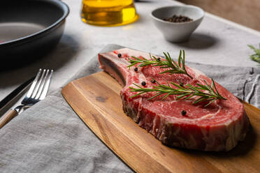 Uncooked t bone beef steak garnished with black pepper and rosemary sprigs placed on wooden chopping board in kitchen - ADSF19270