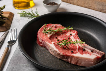 Von oben: rohes Steak mit Knochen, garniert mit Rosmarin und Erbsen mit schwarzem Pfeffer, in einer Pfanne auf dem Küchentisch - ADSF19271