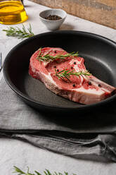 From above uncooked raw t bone steak garnished with rosemary and black pepper peas placed in frying pan on table in kitchen - ADSF19272