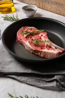 Von oben: rohes Steak mit Knochen, garniert mit Rosmarin und Erbsen mit schwarzem Pfeffer, in einer Pfanne auf dem Küchentisch - ADSF19272