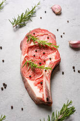 Top view uncooked t bone beef steak garnished with black pepper and rosemary sprigs placed on table in kitchen - ADSF19273