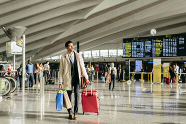 Mann mit dunklem Haar in stilvoller Kleidung geht mit Koffer im Terminal des Flughafens - ADSF19297